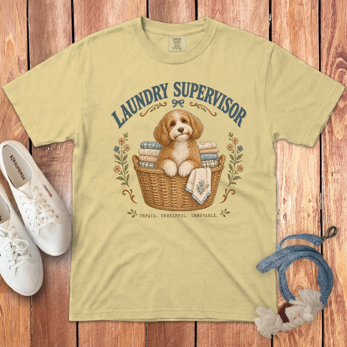 A light yellow t-shirt with a graphic of a fluffy dog sitting in a laundry basket surrounded by flowers and text reading LAUNDRY SUPERVISOR.