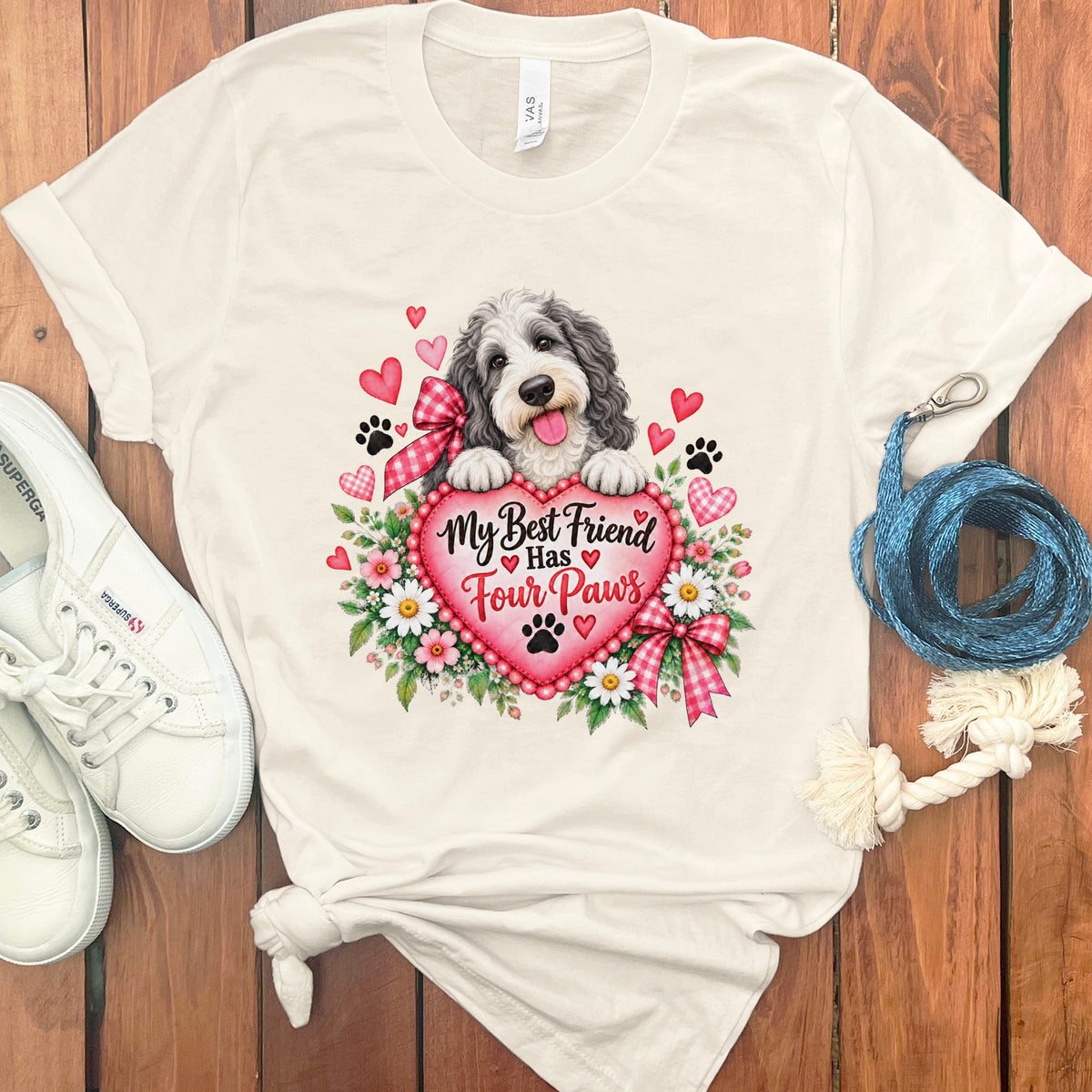 A cream-colored t-shirt features a graphic of a fluffy dog holding a pink heart with the text My Best Friend Has Four Paws, surrounded by flowers, paw prints, and small hearts.