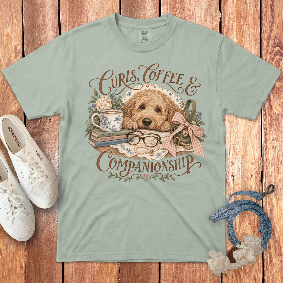 A light green t-shirt featuring a graphic of a labradoodle resting with a coffee cup, books, glasses, and the text Curls, Coffee & Companionship.