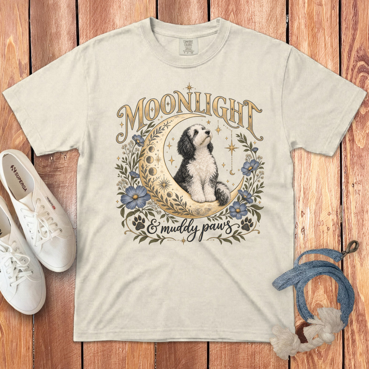 Off-white crewneck t-shirt featuring a vintage illustration of a dog sitting on a crescent moon surrounded by flowers, text reading Moonlight and muddy paws, white sneakers, and a rope leash on a wooden background.