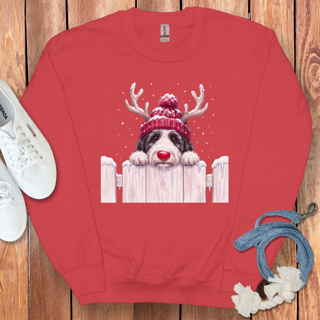 Red crewneck sweatshirt featuring a graphic of a dog wearing reindeer antlers and a knit cap behind a snowy fence.