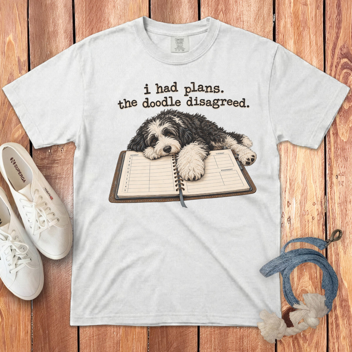 A white t-shirt features a doodle dog resting on an open planner with the text "i had plans. the doodle disagreed."