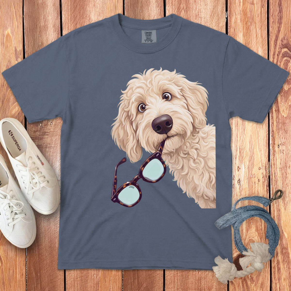 Slate blue t-shirt featuring a doodle dog holding glasses in its mouth, laid on wood with shoes.