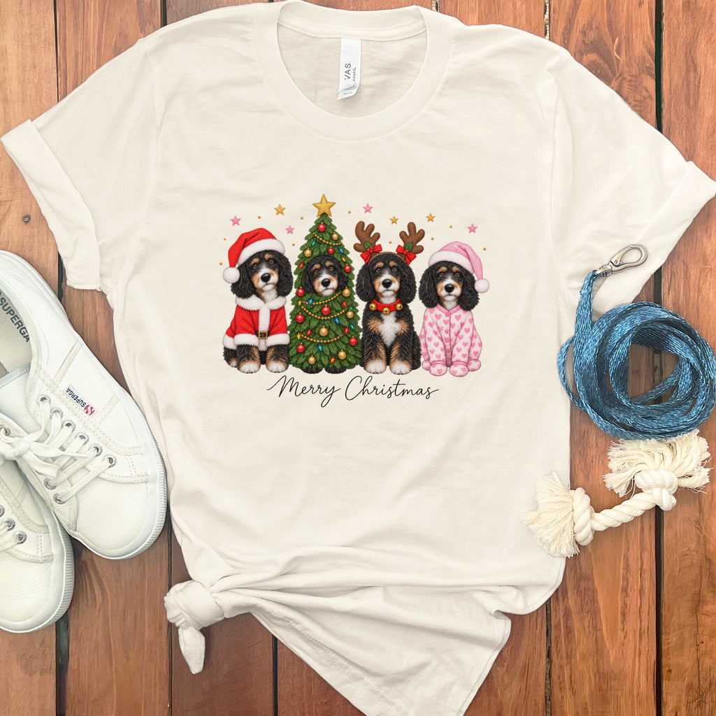 A white t-shirt featuring four Bernedoodles in Christmas attire surrounding a decorated tree, with the text "Merry Christmas" below.