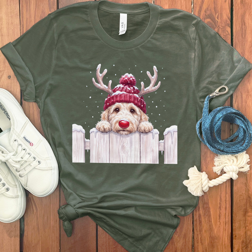 Goldendoodle wearing Santa hat and antlers over white fence, with snow falling.