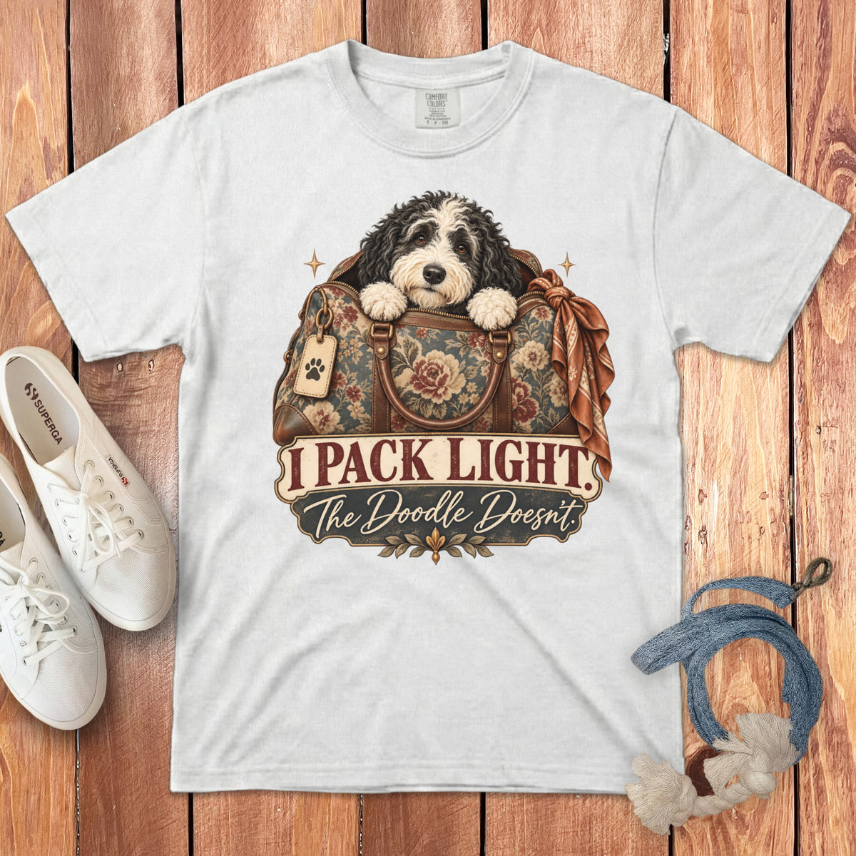 A white crewneck t-shirt featuring a doodle dog inside a floral travel bag with text saying I pack light the doodle doesn't.