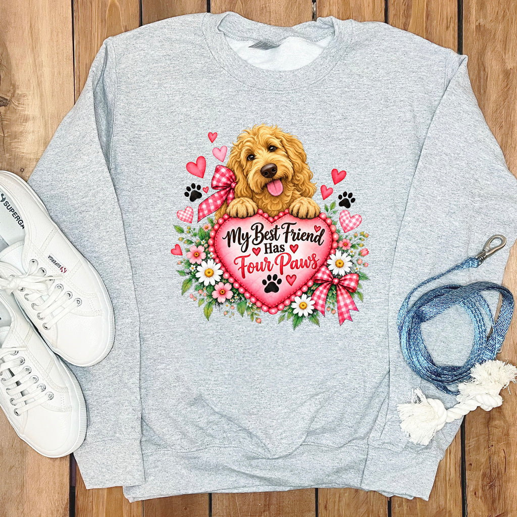 A grey sweatshirt featuring a graphic of a golden doodle dog holding a pink heart that says My Best Friend Has Four Paws, surrounded by flowers, hearts, and paw prints, with white sneakers and a blue leash on a wooden background.