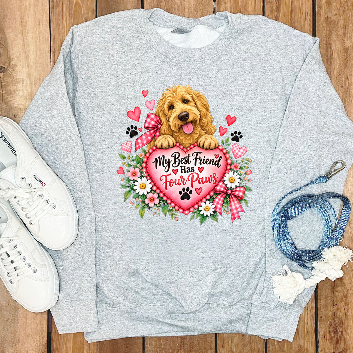 A grey sweatshirt featuring a graphic of a golden doodle dog holding a pink heart that says My Best Friend Has Four Paws, surrounded by flowers, hearts, and paw prints, with white sneakers and a blue leash on a wooden background.