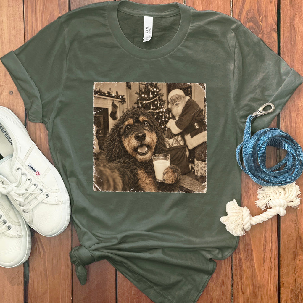 Dark green t-shirt with a vintage graphic of a dog holding a glass of milk and Santa Claus in the background, laid on a wooden surface with white sneakers and a blue leash.