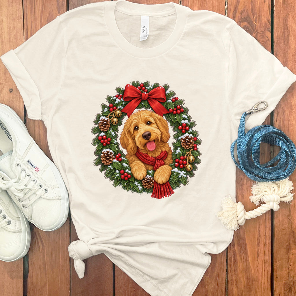A cream-colored t-shirt features a graphic of a golden doodle puppy wearing a red scarf inside a Christmas wreath.