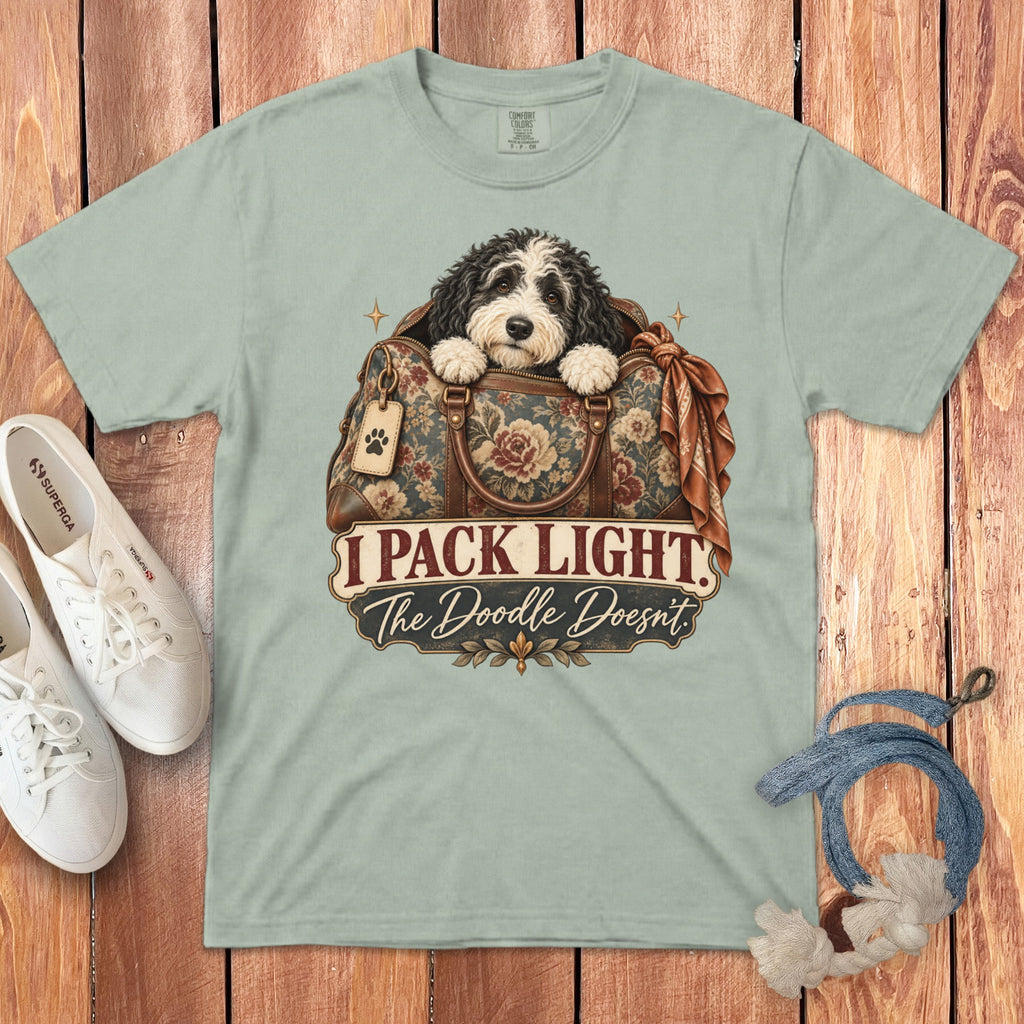 A light green t-shirt displays an illustration of a doodle dog in a floral travel bag with text reading I Pack Light The Doodle Doesnt.