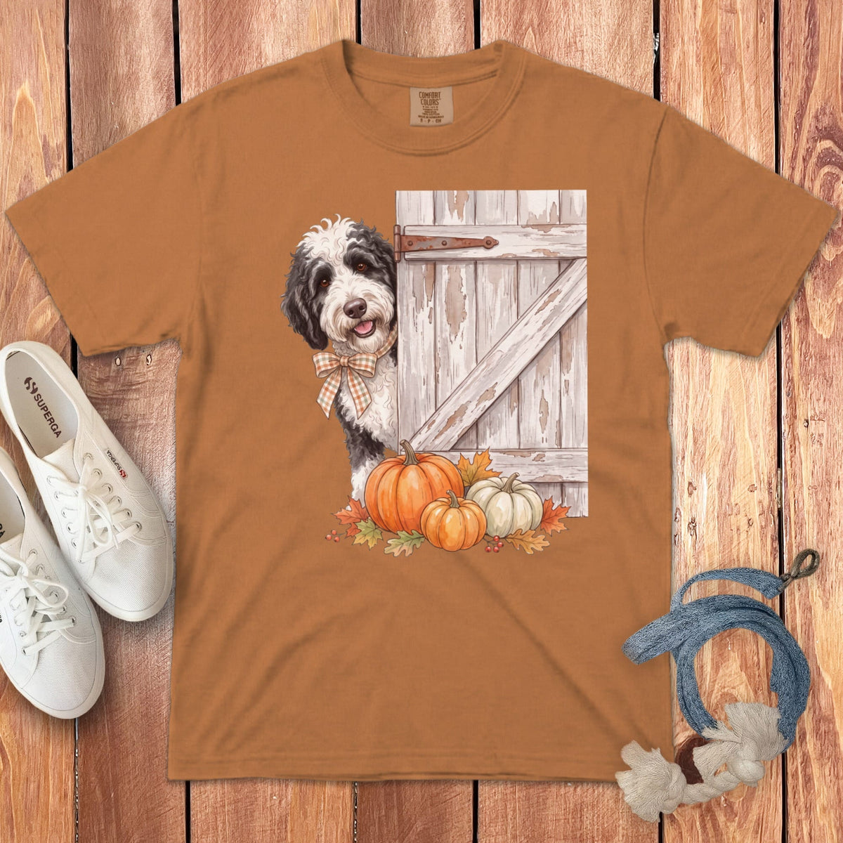 Brown t-shirt with a Bernedoodle peeking from behind a rustic barn door with pumpkins.