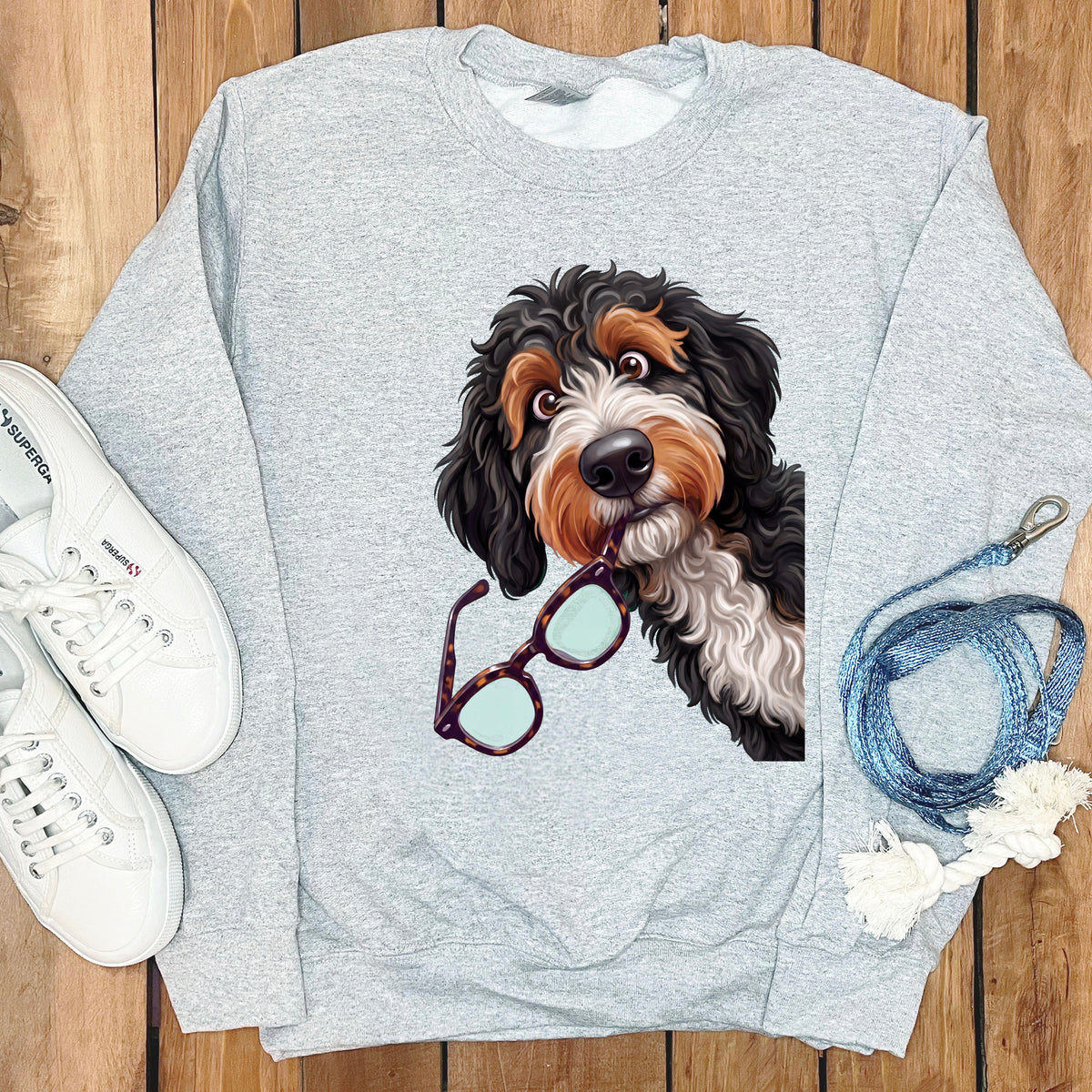Light gray crewneck sweatshirt featuring a graphic of a Bernedoodle holding glasses, with white sneakers and a blue leash.