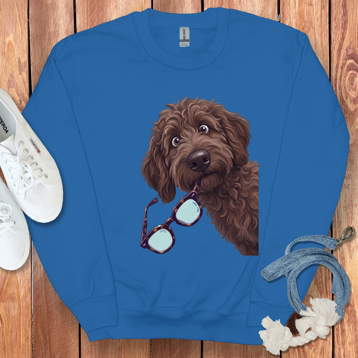 Royal blue sweatshirt featuring a graphic of a brown doodle dog biting tortoiseshell glasses, on wooden background.