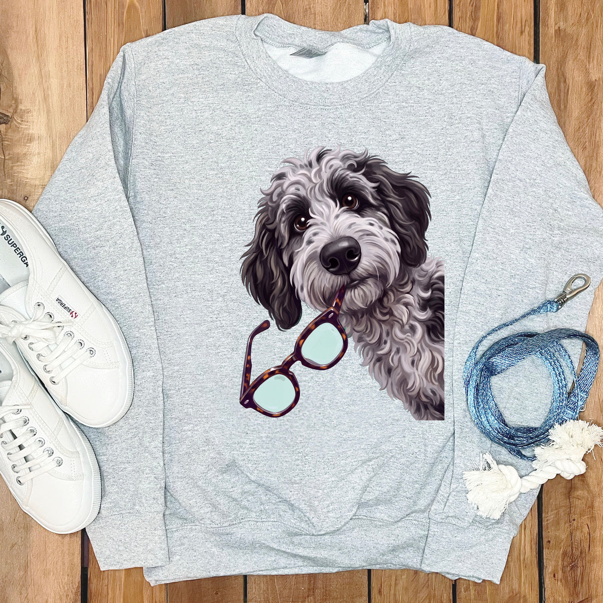 A light gray crewneck sweatshirt featuring a graphic of a black and white doodle dog holding glasses in its mouth.