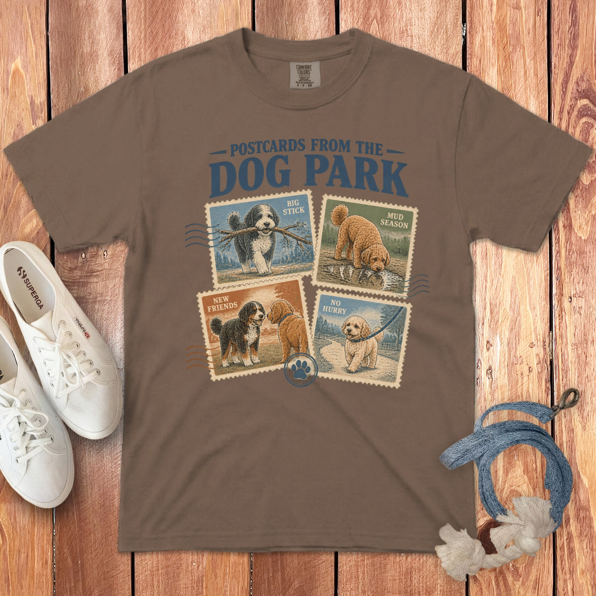 Brown t-shirt featuring graphic text Postcards from the Dog Park above four vintage-style dog postage stamp illustrations.