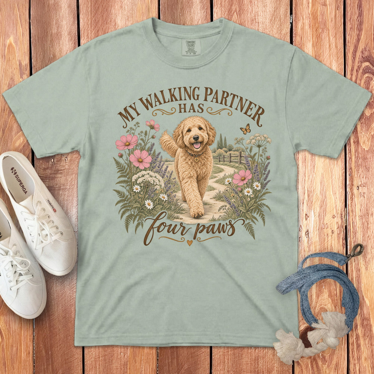 A light green t-shirt with a graphic of a golden doodle dog walking among wildflowers.