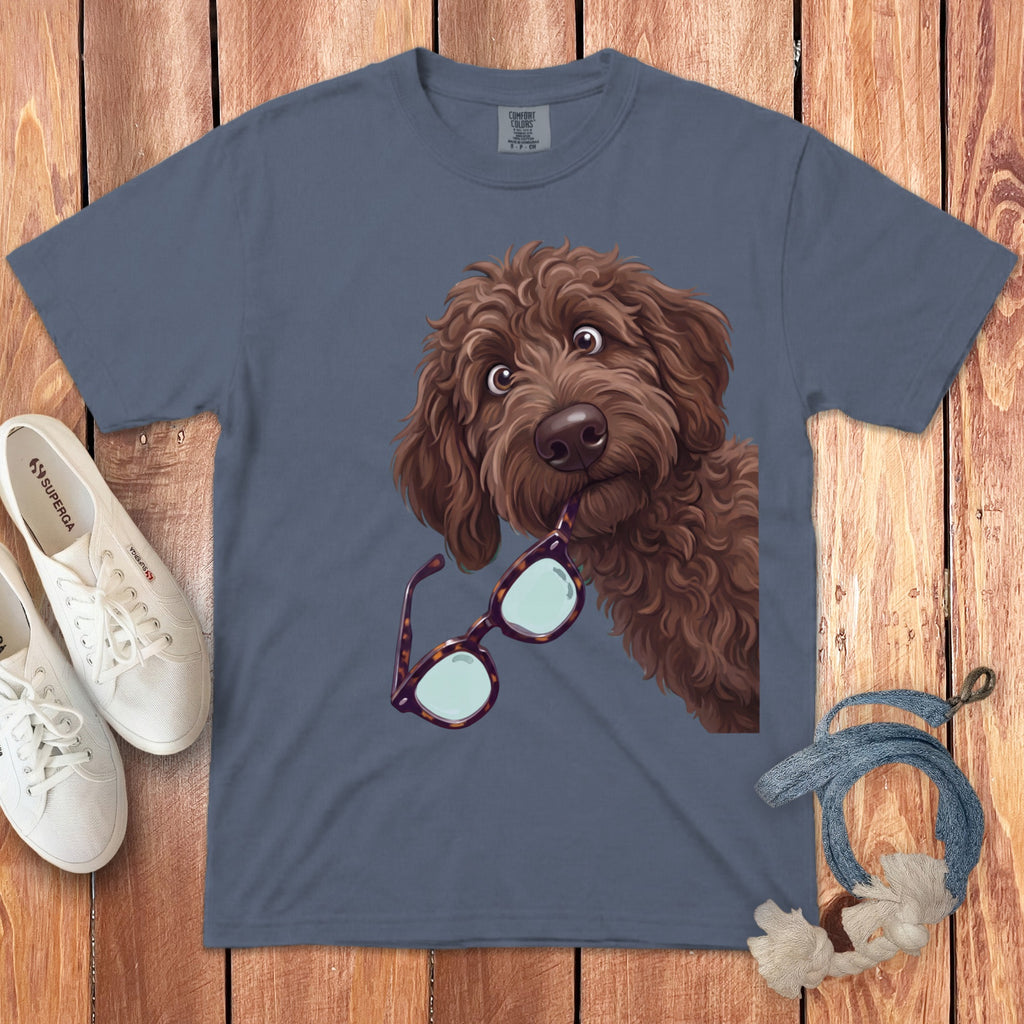 A slate blue crewneck t-shirt featuring a brown doodle dog holding glasses in its mouth, laid on a wooden background with white shoes and a lanyard.