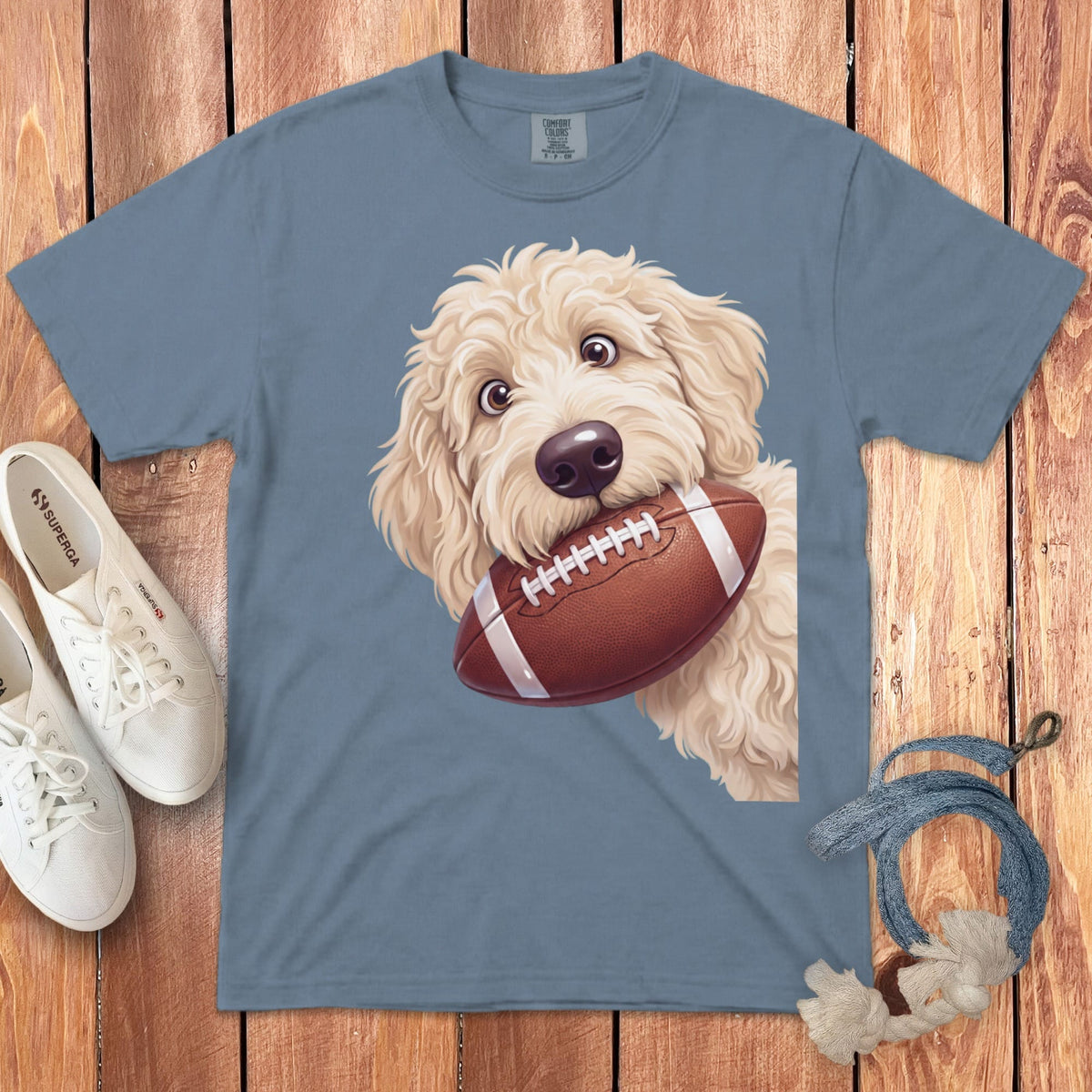 Goldendoodle puppy with a football in its mouth on a blue t-shirt.