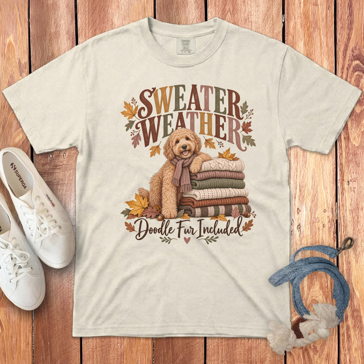 An off-white t-shirt featuring a cartoon doodle dog, stacked sweaters, autumn leaves, and sweater weather text.