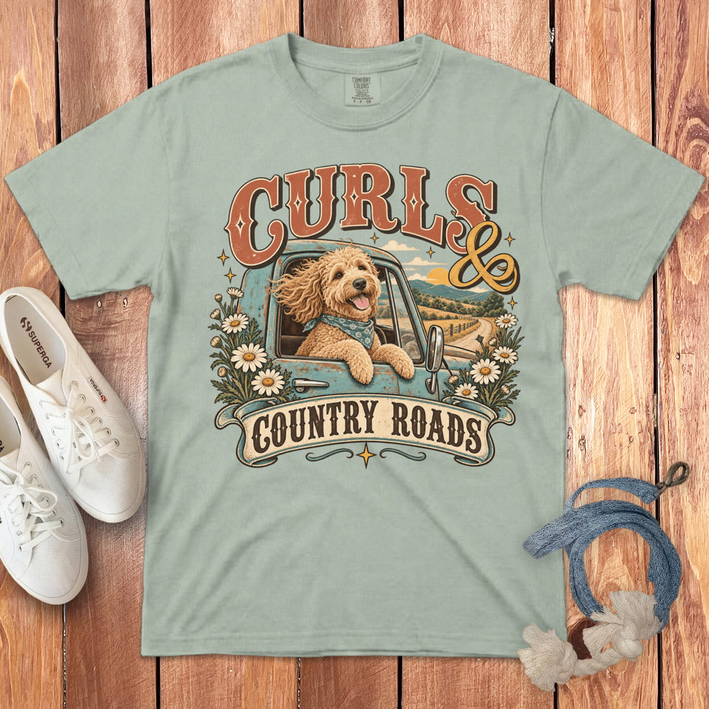 A sage green t-shirt featuring a graphic of a curly doodle dog in a truck with daisies and the text Curls and Country Roads.