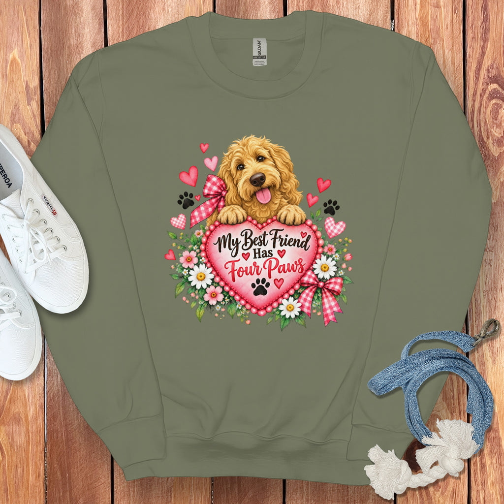 An olive green crewneck sweatshirt featuring a graphic of a fluffy gold doodle puppy resting on a pink heart inscribed with the text My Best Friend Has Four Paws, surrounded by flowers, paw prints, and smaller hearts, set against a wooden background with white sneakers and a blue leash.