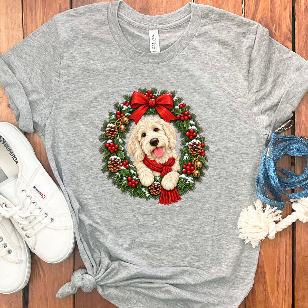 Heather grey t-shirt featuring a goldendoodle wearing a red scarf inside a Christmas wreath, accompanied by white sneakers and a blue leash.