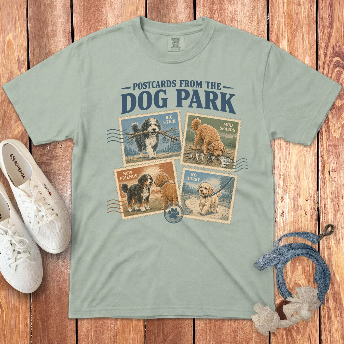 Sage green t-shirt with Postcards From The Dog Park graphic featuring four dog stamp illustrations, surrounded by white shoes and a dog leash on wood.