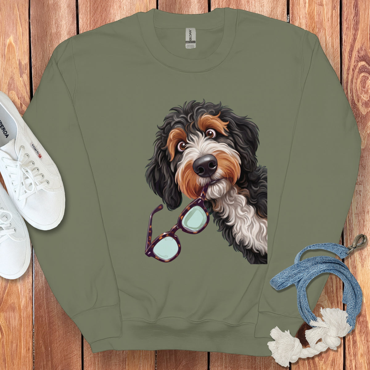 An olive green crewneck sweatshirt featuring an illustration of a doodle dog holding glasses in its mouth.