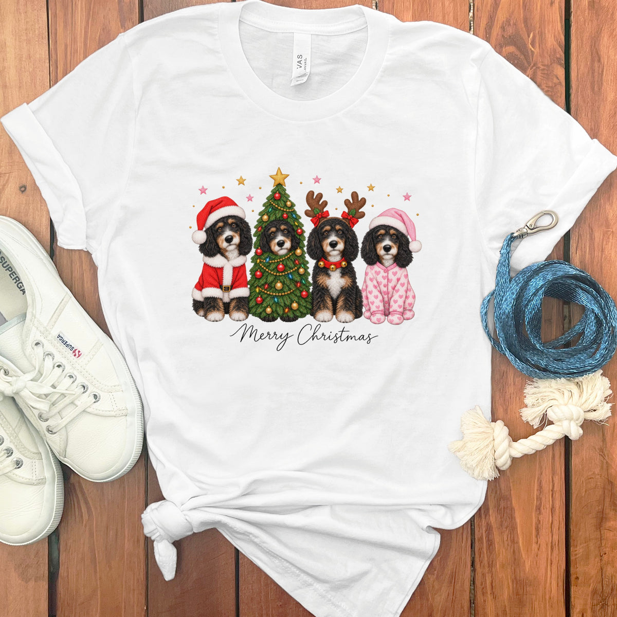 A white t-shirt featuring a graphic of four Bernedoodles in Christmas attire surrounding a decorated tree, with Merry Christmas text below.