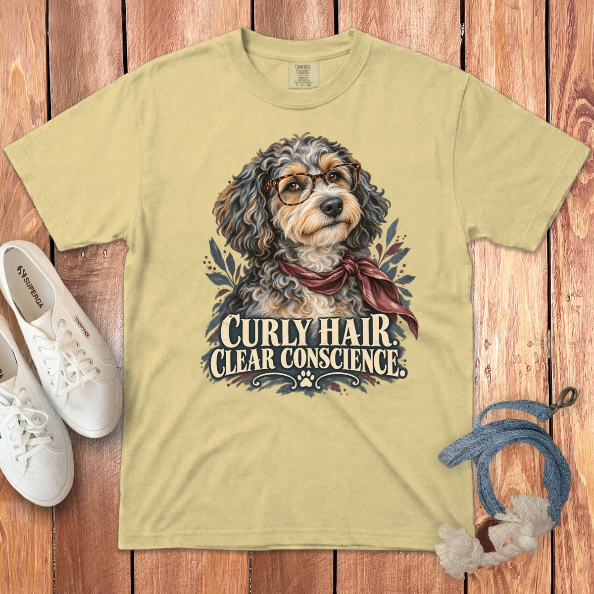 A pale yellow t-shirt features a graphic of a fluffy dog wearing glasses and a scarf, accompanied by the text "Curly Hair Clear Conscience."