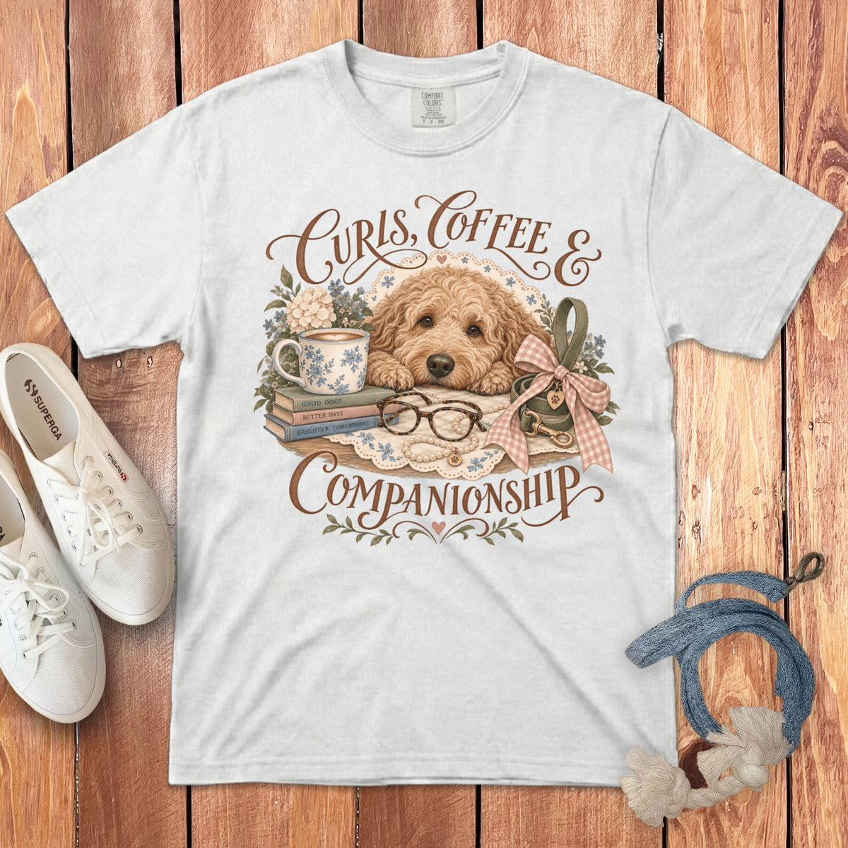 A white t-shirt featuring a graphic of a golden doodle dog, books, a coffee mug, and text reading Curls, Coffee & Companionship.