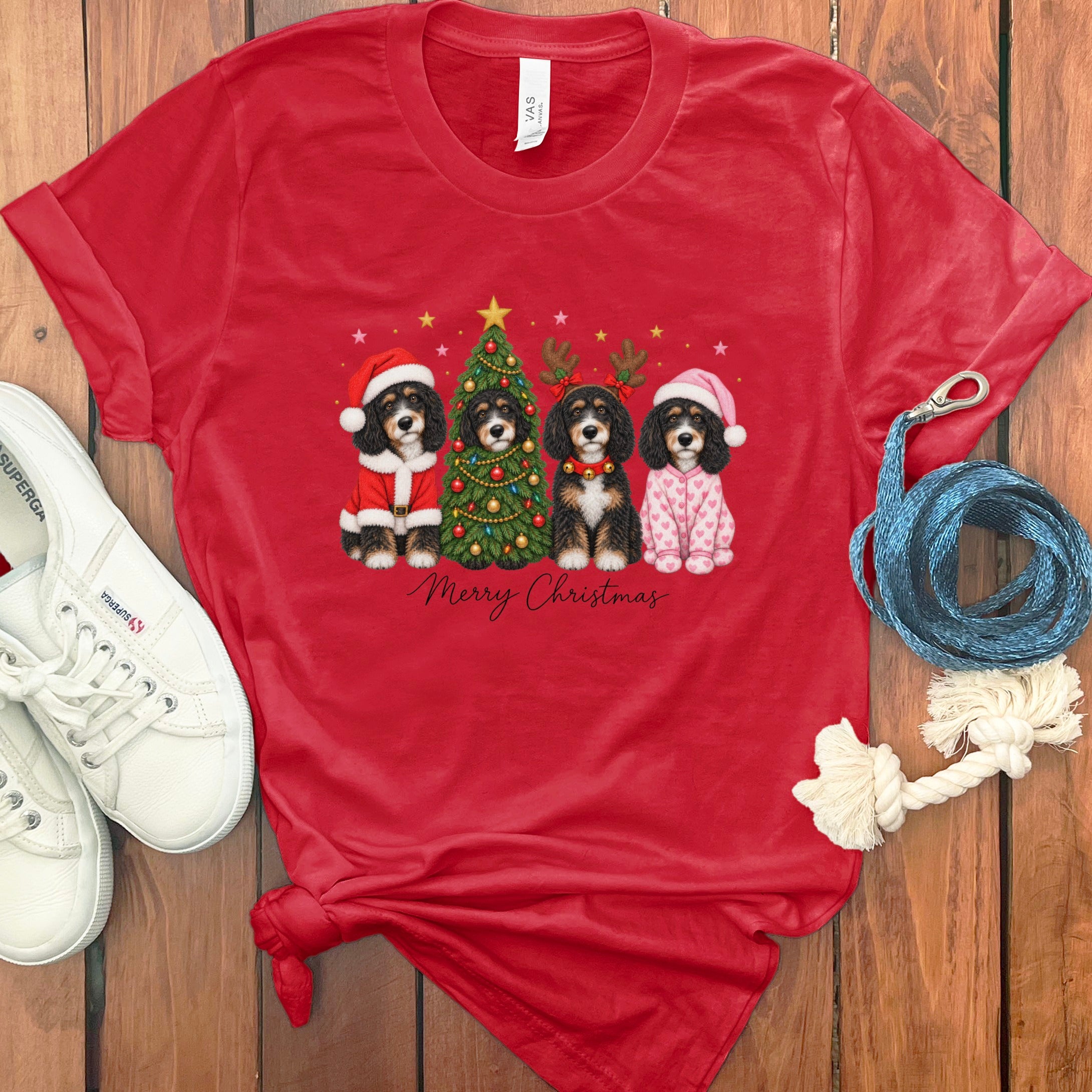 A red t-shirt with a graphic of four Bernedoodles in Christmas attire surrounding a decorated tree, accompanied by white sneakers and a blue leash.