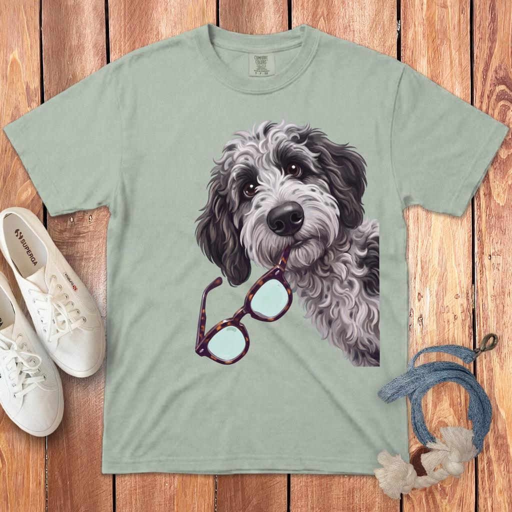 A sage green t-shirt featuring a graphic of a fluffy dog holding glasses in its mouth, laid flat on a wooden surface with white sneakers and a blue rope accessory.