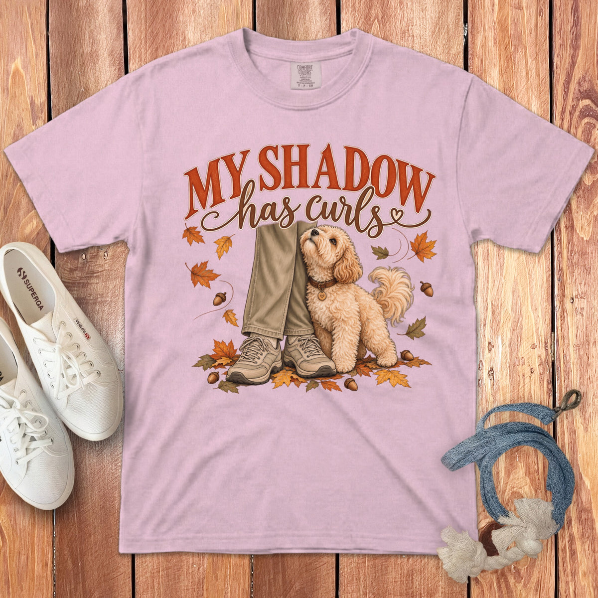 A light pink t-shirt with an illustration of a fluffy dog sitting beside someone's legs, surrounded by autumn leaves and acorns, and the text "MY SHADOW has curls".