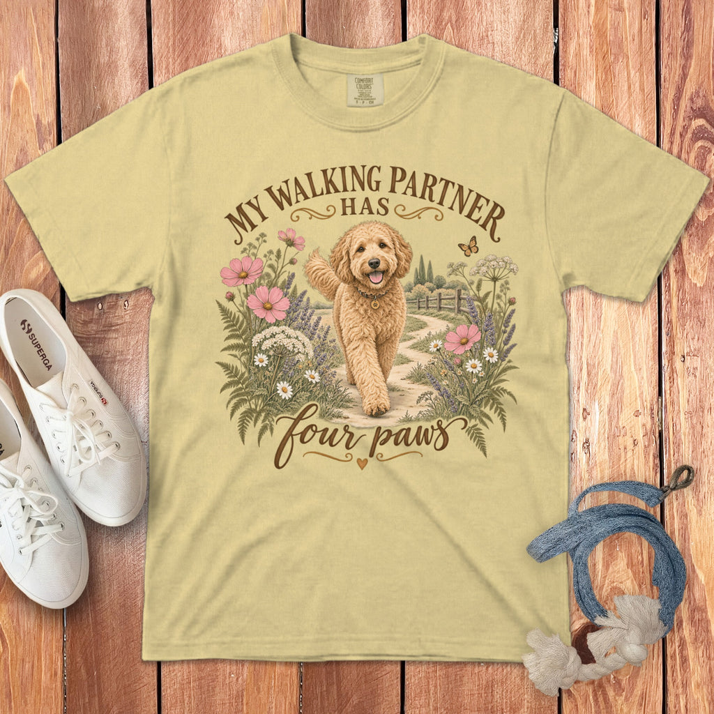 A light yellow t-shirt with a graphic of a golden doodle dog walking among flowers and the text "MY WALKING PARTNER HAS FOUR PAWS," laid on wood alongside white sneakers and a blue leash.