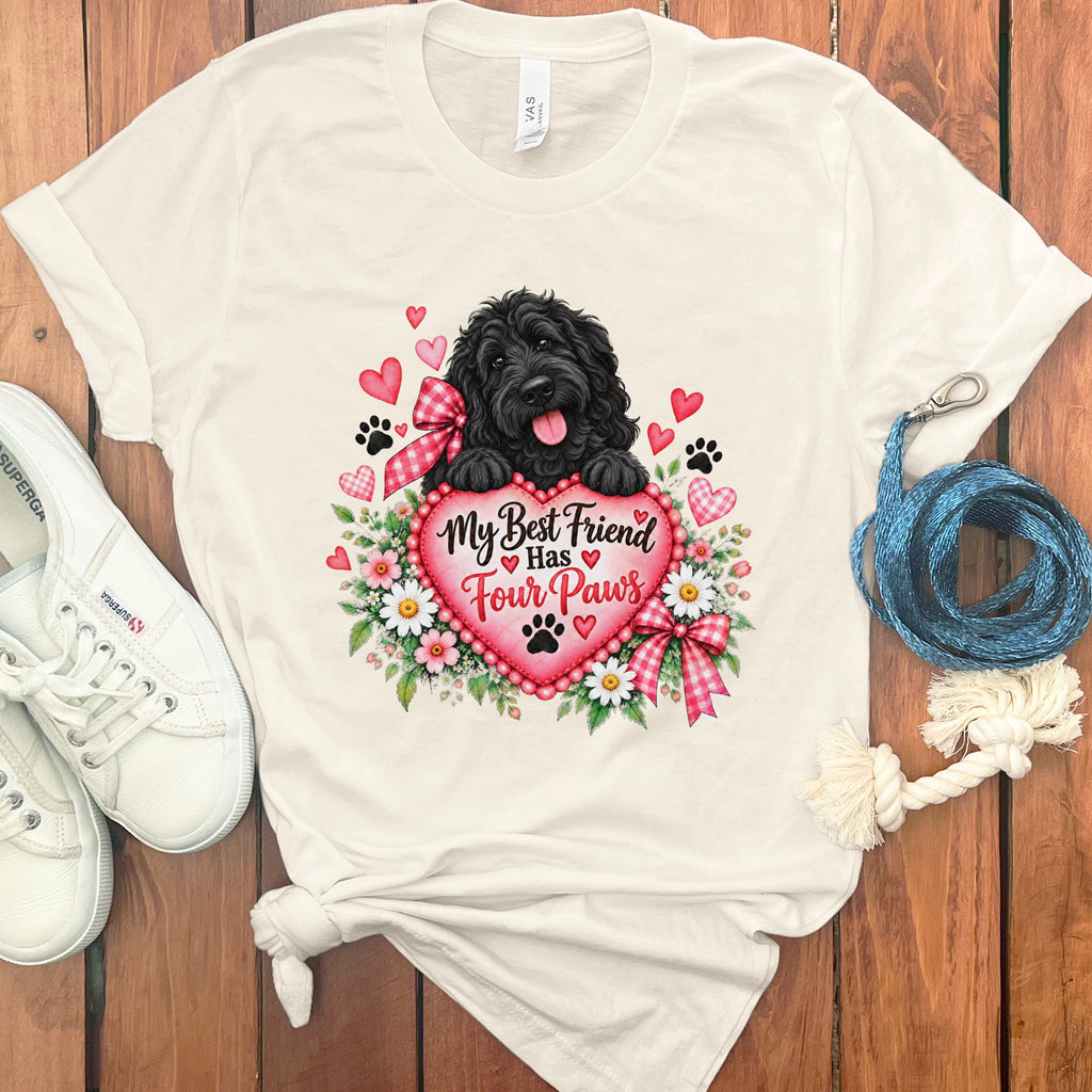 Off-white t-shirt with a graphic of a black dog holding a pink heart reading My Best Friend Has Four Paws, surrounded by flowers, hearts, and paw prints, flanked by white sneakers and a blue leash on a wooden surface.
