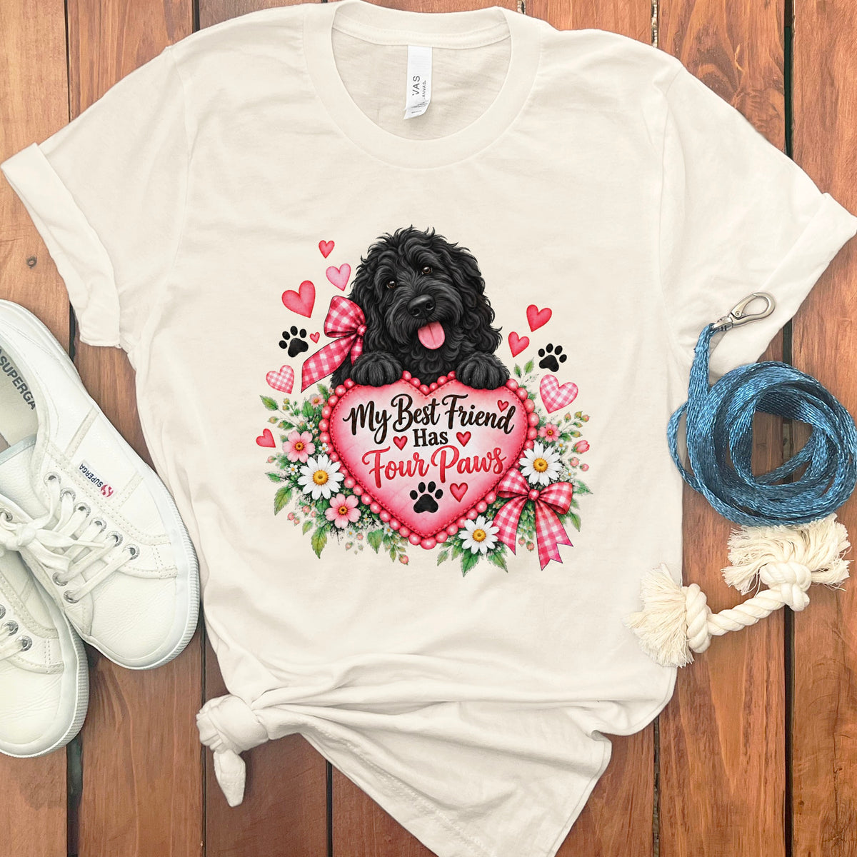 Off-white t-shirt with a graphic of a black dog holding a pink heart reading My Best Friend Has Four Paws, surrounded by flowers, hearts, and paw prints, flanked by white sneakers and a blue leash on a wooden surface.