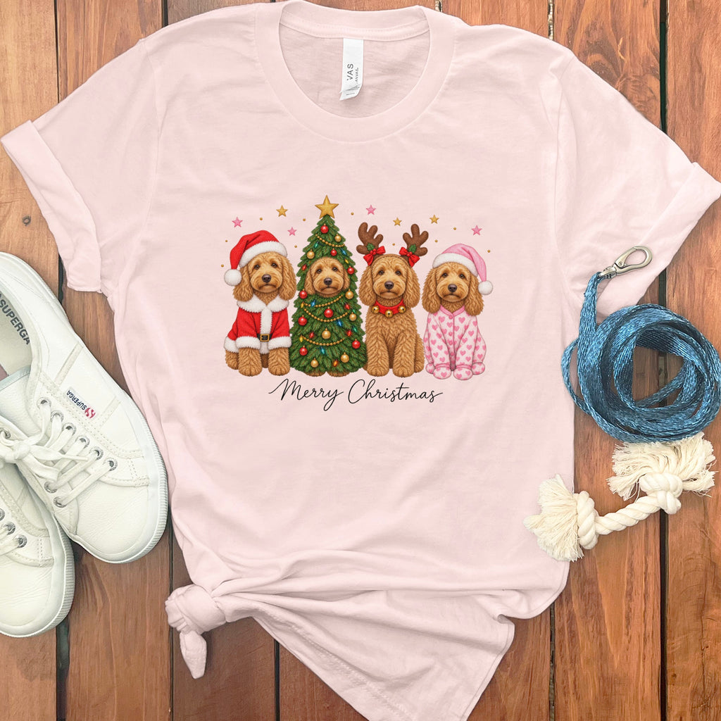 A light pink t-shirt with a graphic of four doodle dogs celebrating Christmas, surrounded by white shoes and a blue leash on a wooden background.