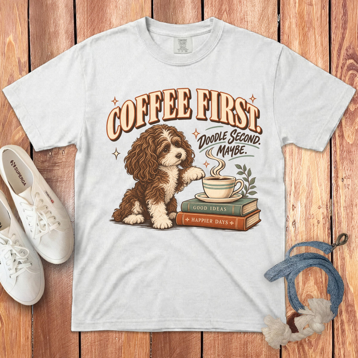 A white t-shirt featuring a graphic of a brown doodle dog beside a coffee cup on books, with text reading COFFEE FIRST DOODLE SECOND MAYBE.