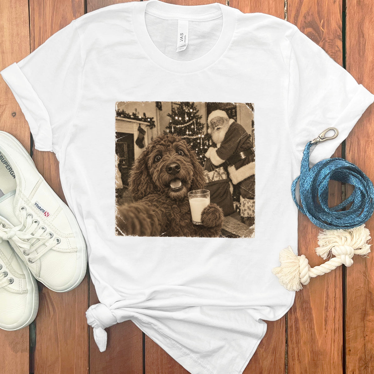 A white t-shirt featuring a sepia-toned graphic of a fluffy brown dog holding a glass of milk in front of Santa Claus.