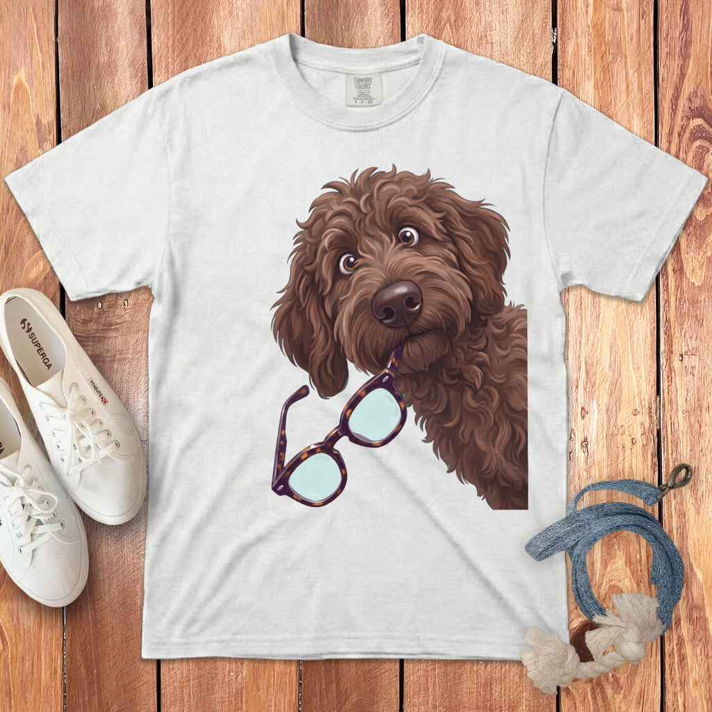 A white t-shirt featuring a cartoon illustration of a brown doodle dog holding glasses in its mouth.