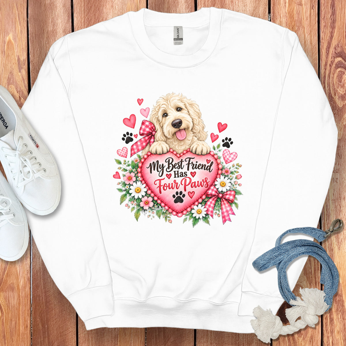 A white crewneck sweatshirt features a graphic of a golden puppy resting on a pink heart inscribed with "My Best Friend Has Four Paws," surrounded by flowers, hearts, and paw prints, laid on a wooden background with white shoes and a blue leash.
