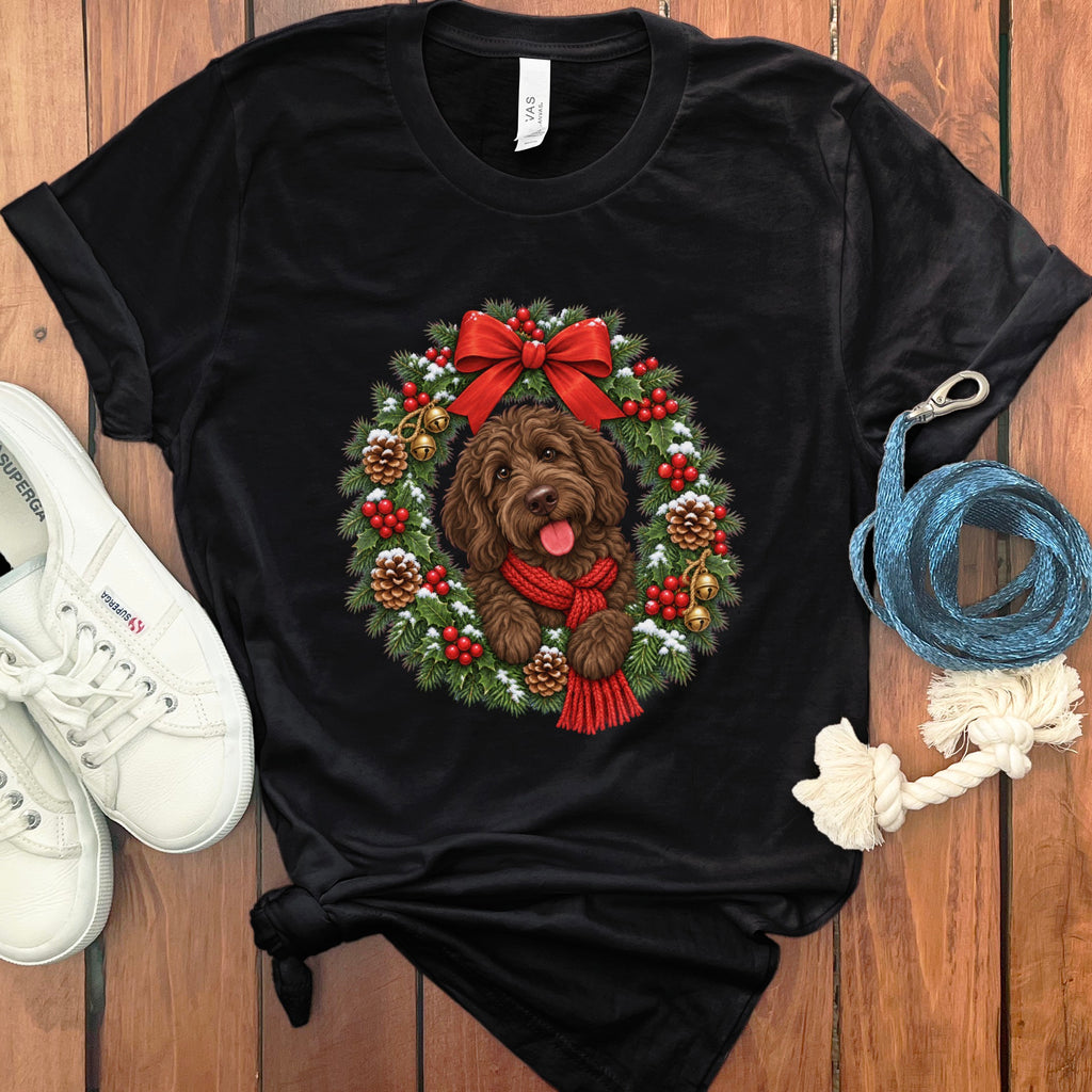 A black t-shirt features a graphic of a brown doodle dog wearing a red scarf inside a Christmas wreath, laid out on a wooden surface with white sneakers and a blue leash.