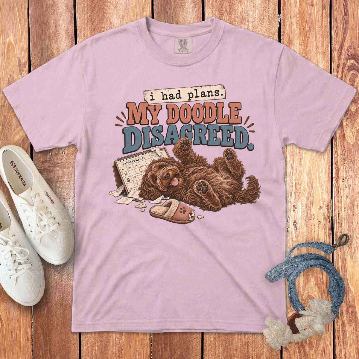 Light pink t-shirt featuring a cartoon of a doodle dog lying on its back next to a chewed calendar and slipper.