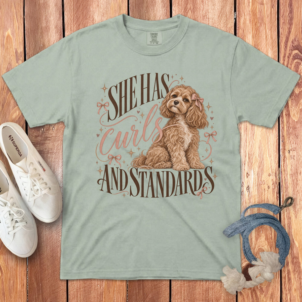 A sage green t-shirt with a graphic of a curly brown dog and the text She Has Curls And Standards.