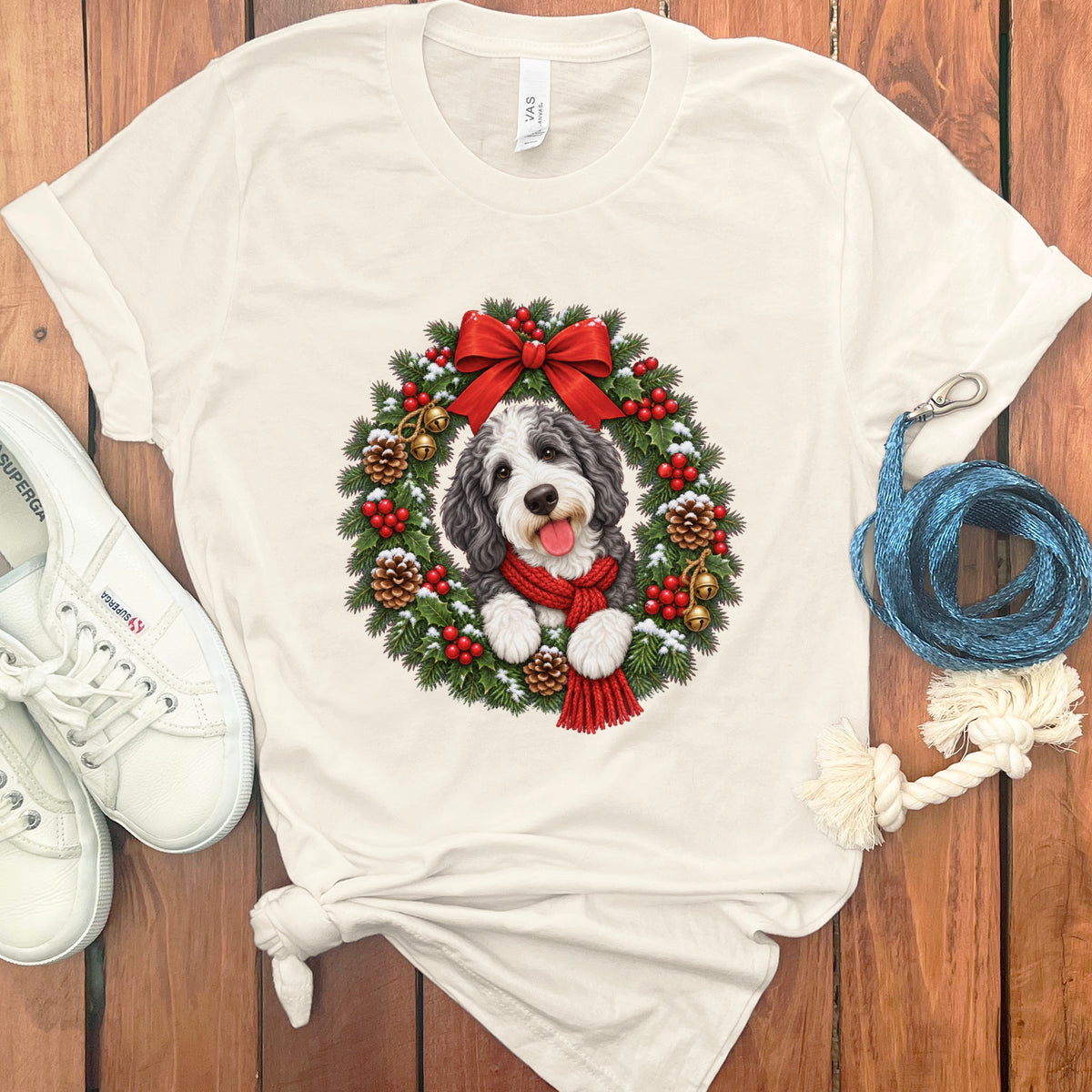 Beige t-shirt featuring a doodle dog wearing a red scarf inside a Christmas wreath on a wooden surface.