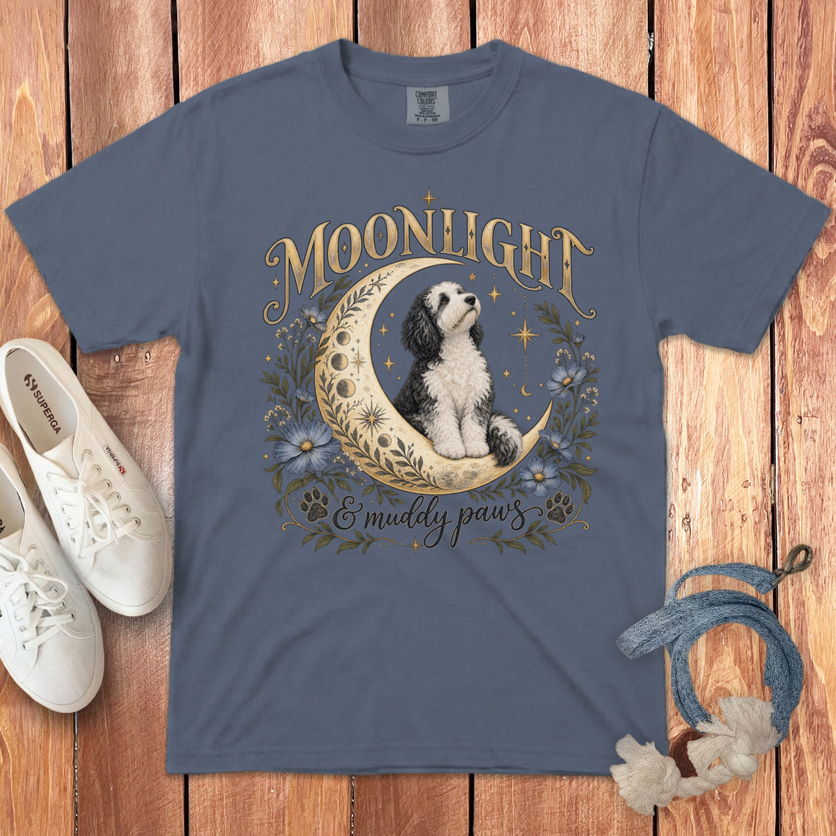 Blue t-shirt featuring a graphic of a black and white dog sitting on a crescent moon with flowers and the text Moonlight & muddy paws.