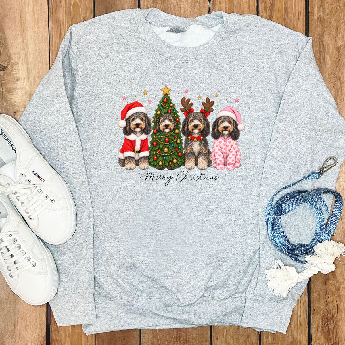 Light gray crewneck sweatshirt featuring a graphic of four doodle dogs celebrating Christmas, surrounded by white sneakers and a blue leash.