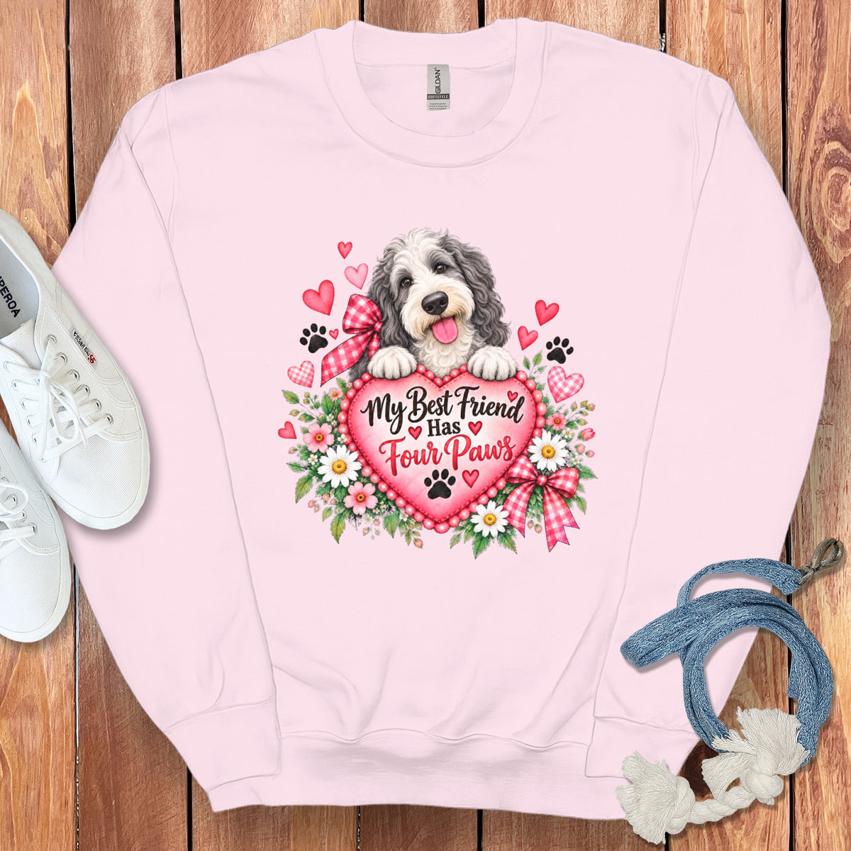 Light pink crewneck sweatshirt featuring a graphic of a fluffy dog resting on a heart with text reading My Best Friend Has Four Paws surrounded by flowers and paw prints, displayed flat on a wooden surface with white sneakers and a blue leash.
